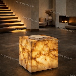 May include: A square, illuminated onyx side table with a book on top. The table has a warm, amber glow with brown veining. The background includes a staircase, fireplace, and seating area, creating a cozy ambiance.