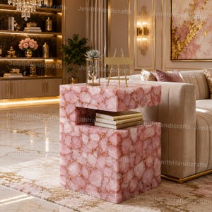 May include: A pink side table with a unique design. The table is made of pink stone pieces with gold accents. It has a two-tiered design with a shelf for storage. A small gold decorative piece and a glass vase with a plant are on top.