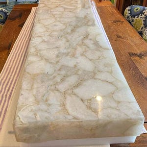 Puede incluir: Una encimera rectangular con una superficie pulida. La encimera presenta un patrón de formas orgánicas blanquecinas y bronceadas, que se asemejan a la piedra natural. Los bordes son lisos y la pieza descansa sobre espuma blanca.