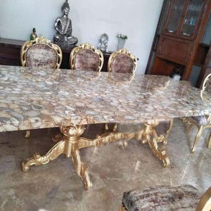 Puede incluir: Una mesa de comedor grande con una tapa única de piedra de ágata y patas pintadas de oro. La mesa está rodeada de seis sillas con respaldos ornamentados y tapicería de felpa.