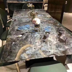 Puede incluir: Una gran mesa rectangular de comedor con una tapa de mármol azul y gris. La mesa tiene una base negra y patas doradas. Hay varios elementos decorativos en la mesa, incluyendo un jarrón de flores blancas, un candelero dorado y una escultura de vidrio azul.