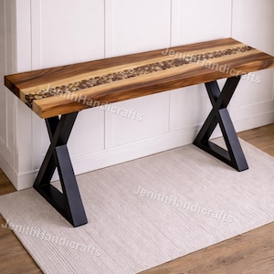 May include: A handcrafted wooden bench with a unique design. The bench features a dark wood top with a central inlay of lighter wood and decorative elements. It rests on black metal X-shaped legs. Suitable for indoor use.