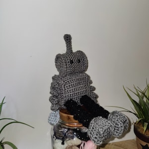Può includere: Un giocattolo robot di uncinetto grigio con gambe nere seduto su un barattolo di vetro pieno di filato. Il robot ha una piccola antenna sulla testa e occhi neri.