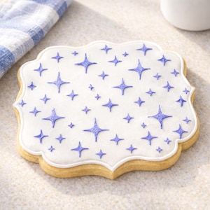 Pode incluir: Um biscoito decorado com uma base de glacê branco e um padrão de estrelas azuis. O biscoito tem uma borda ondulada e uma aparência marrom clara, assada. Um pano listrado azul e branco está no fundo.