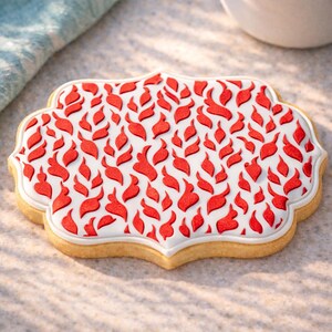 May include: A decorated sugar cookie with a white base and red leaf-like designs. The cookie has a scalloped edge and a white border. The cookie is on a light-colored surface.