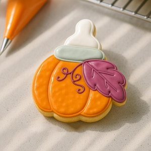May include: A decorated pumpkin-shaped cookie with orange icing, a white top, and a purple leaf. The cookie has a light brown base and is decorated with intricate details, perfect for fall or Thanksgiving.