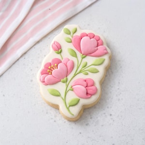 Peut inclure: Un biscuit décoré avec un motif floral. Le biscuit présente des fleurs roses avec des feuilles et des tiges vertes sur un fond blanc. Le biscuit est bordé d'un glaçage blanc et a une base de biscuit brun clair.