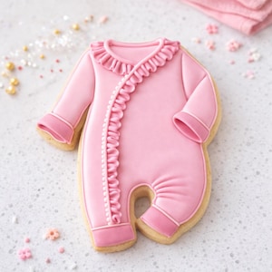 May include: A pink baby onesie cookie with a ruffled collar and trim. The cookie is decorated with pink icing and has a light-colored cookie base. The onesie is a light pink color with darker pink accents. The cookie is decorated with small pink and gold sprinkles.