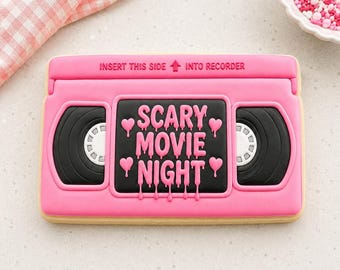 May include: A rectangular biscuit shaped like a pink VHS tape with black reels and a central label reading SCARY MOVIE NIGHT in a dripping font. The top edge features the embossed text INSERT THIS SIDE INTO RECORDER with an upward arrow.