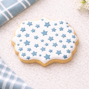 May include: A star-shaped cookie with a white icing design and blue star imprints. The cookie has a light brown, buttery base and a scalloped edge. The cookie is on a white surface with a blue and white checkered cloth in the background.