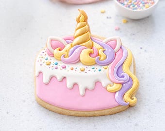 May include: A unicorn-shaped cookie with a pink base, white icing, and colorful sprinkles. The cookie features a golden horn, pink and yellow ears, and flowing purple and yellow hair. A bowl of sprinkles is in the background.