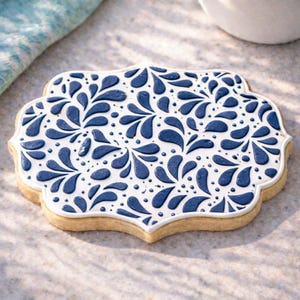 Puede incluir: Una galleta decorada con una base blanca y un patrón floral azul. La galleta tiene un borde festoneado y se coloca sobre una superficie de color claro. El diseño azul está formado por formas de gota y puntos.