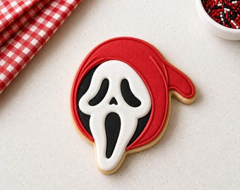 May include: A sugar biscuit shaped like a ghost face mask, decorated with smooth red icing for the hood and white icing for the face, featuring black icing details for the eyes and mouth, presented as a single baked treat on a flat surface.