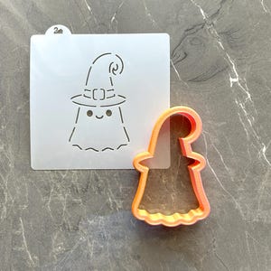May include: A translucent square stencil featuring a cutout design of a ghost wearing a witch hat with a small number 2 tab at the top, paired with an orange plastic cookie cutter shaped like the same ghost figure with a curved witch hat.