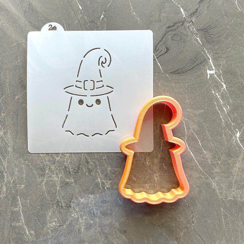 May include: A translucent square stencil featuring a cutout design of a ghost wearing a witch hat with a small number 2 tab at the top, paired with an orange plastic cookie cutter shaped like the same ghost figure with a curved witch hat.