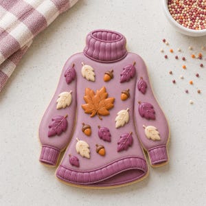 Puede incluir: Una galleta con forma de suéter decorada con glaseado morado, que presenta un cuello de tortuga y puños acanalados. La superficie está adornada con pequeñas decoraciones de glaseado en relieve de hojas de arce naranjas, hojas color crema, hojas moradas y bellotas marrones.