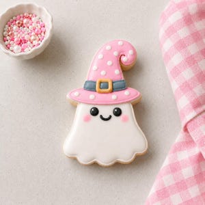 May include: A ghost-shaped sugar biscuit decorated with white icing and a pink witch hat featuring white polka dots, a blue band, and a yellow buckle. The ghost has black eyes, a small black smile, and soft pink cheeks, accompanied by pink sprinkles.
