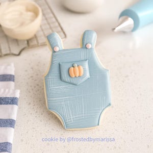 May include: A light blue onesie-shaped cookie with a small orange pumpkin on the pocket. The cookie has a textured pattern and is decorated with light blue icing. The text "cookie by @frostedbymarissa" is at the bottom.