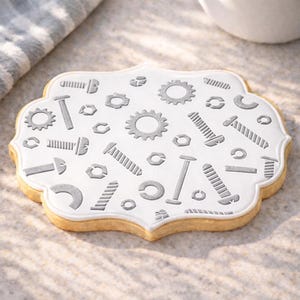 May include: A decorated cookie with a white icing design featuring gray illustrations of gears, bolts, screws, and nuts. The cookie has a light brown, scalloped edge and sits on a light-colored surface.