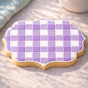 May include: A decorative cookie with a purple and white checkered pattern. The cookie has a unique, scalloped shape and a white border. The cookie is light brown around the edges.