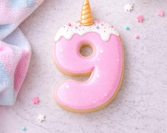 May include: A pink number nine cookie decorated with white icing, sprinkles, and a gold unicorn horn. The cookie is on a light gray surface with scattered star-shaped sprinkles and a pastel-colored cloth.