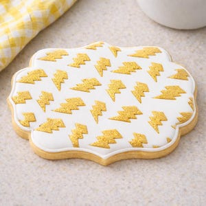 May include: A decorated sugar cookie with a white icing base and gold glitter lightning bolt designs. The cookie has a scalloped edge and a light brown base. A yellow and white checkered cloth is in the background.