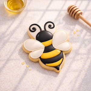 May include: A decorated bee-shaped cookie with black, yellow, and white icing. The cookie has a detailed design, including wings and stripes. A small bowl of honey and a wooden honey dipper are in the background.