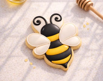 May include: A decorated bee-shaped cookie with black, yellow, and white icing. The cookie has a detailed design, including wings and stripes. A small bowl of honey and a wooden honey dipper are in the background.