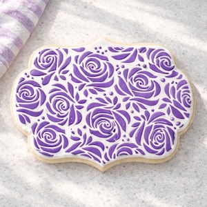 May include: A decorated sugar cookie with a white base and a purple floral design. The cookie has a scalloped edge and is decorated with a pattern of roses and leaves. A purple and white striped cloth is in the background.