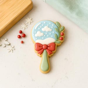 May include: A decorated sugar cookie in the shape of a snow globe. The cookie features a light blue sky with white clouds and snow, a red bow, and green leaves with red berries. The cookie is on a white surface.