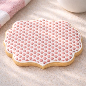 May include: A decorated sugar cookie with a scalloped edge. The cookie has a white base with a pattern of pink dots. A white border outlines the cookie. A pink and white striped cloth is in the background.