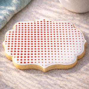 May include: A decorative cookie with a white icing design featuring a pattern of red dots. The cookie has a light brown, scalloped edge and is shaped like a stylized quatrefoil. The cookie is a sweet treat.