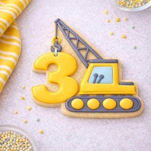 May include: A decorated sugar cookie in the shape of a yellow construction crane with the number 3. The crane has a gray arm and a yellow cab with a blue window. The cookie is on a light-colored surface with sprinkles.