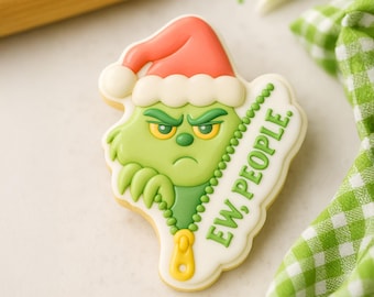 May include: A Christmas cookie shaped like a green character with a red Santa hat and the text "EW, PEOPLE." The cookie has a yellow zipper detail and is decorated with white icing.