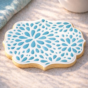 May include: A decorative sugar cookie with a white and blue floral design. The cookie has a light brown base with a white border and a blue floral pattern. The cookie is shaped like a decorative plaque.
