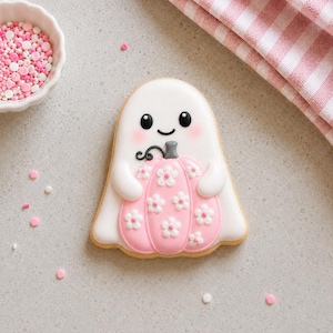 Könnte beinhalten: Ein geisterförmiger Keks, verziert mit weißem Zuckerguss, schwarzen Augen, einem kleinen Lächeln und rosa Wangen. Der Geist hält einen hellrosa Kürbis mit weißen Blumenverzierungen, einem grauen Stiel und einer schwarzen Ranke, neben rosa Streuseln.