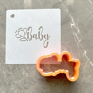 Puede incluir: Un est&eacute;ncil blanco con la palabra "Baby" recortada en una fuente cursiva, acompa&ntilde;ada de un dise&ntilde;o floral. Un cortador de galletas naranja con la forma de la palabra "Baby" est&aacute; al lado del est&eacute;ncil. El est&eacute;ncil tiene un peque&ntilde;o recorte en forma de coraz&oacute;n.