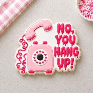May include: A sugar biscuit shaped like a pink rotary telephone with a coiled cord, featuring the text NO YOU HANG UP! in dripping hot pink icing. The dial is decorated with small red hearts surrounding a black centre on a white icing base.