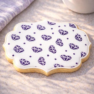 May include: A sugar cookie with a white icing top decorated with purple diamond shapes and dots. The cookie has a scalloped edge and a golden-brown base. The cookie is on a light-colored surface.