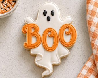 May include: A ghost-shaped sugar cookie decorated with smooth white icing and black icing for the eyes and mouth. The word BOO is written across the center in raised orange icing. The cookie has a wavy, flowing bottom edge and a rounded top.
