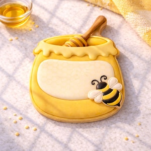 May include: A yellow honey pot-shaped cookie with a wooden honey dipper inside. The cookie has a white honeycomb design and a black and yellow bee decoration. The cookie is surrounded by small yellow sprinkles.