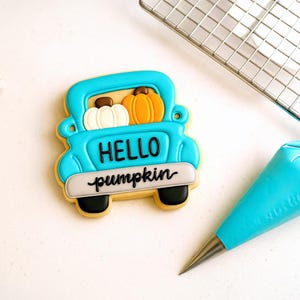 Może przedstawiać: Ozdobione ciastko w kształcie jasnoniebieskiej ciężarówki z dwiema dyniami w skrzyni. Ciastko ma napis "HELLO pumpkin". Dynie są białe i pomarańczowe. Ciastko jest ozdobione czarnym, białym i beżowym lukrem.