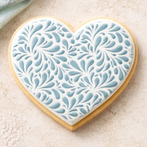 May include: A heart-shaped cookie with a white icing design featuring a blue floral pattern. The cookie has a light brown, golden-baked edge. The design is a delicate, elegant style.