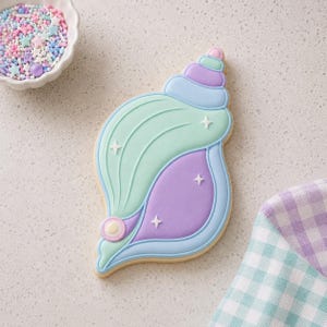 Puede incluir: Una galleta de azúcar con forma de concha marina decorada con glaseado pastel liso en tonos verde menta, lavanda y azul claro. La galleta presenta detalles de glaseado en relieve, pequeñas estrellas blancas y un adorno circular perlado.