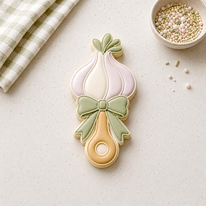 May include: A garlic-shaped sugar cookie with a green bow and yellow handle. The cookie is decorated in light pink, white, and green icing. A small bowl of sprinkles and a green and white checkered cloth are in the background. The cookie is a sweet treat.