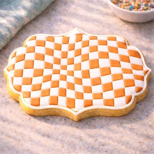 May include: A decorated cookie with a white and orange checkered pattern. The cookie has a decorative shape with a white border and a light brown base. The cookie is on a light-colored surface.
