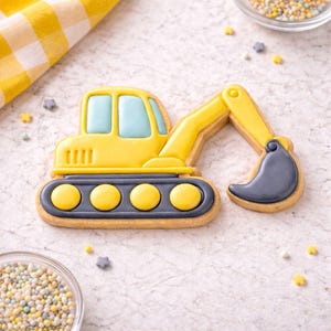 Puede incluir: Una galleta amarilla con forma de excavadora con detalles de ventanas azules y un cucharón gris. La galleta tiene una base gris con círculos amarillos y está decorada con glaseado amarillo. La galleta está sobre una superficie blanca con chispas.