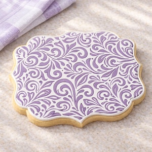 May include: A decorative cookie with a white border and a lavender floral design. The cookie has a light brown base and a scalloped edge. A lavender and white checkered cloth is in the background.
