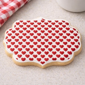 Può includere: Un biscotto a forma di cuore con un bordo di glassa bianca e un motivo di cuori rossi. Il biscotto ha un aspetto marrone chiaro, cotto ed è posto su uno sfondo neutro. Un panno a scacchi rosso e bianco è visibile sullo sfondo.