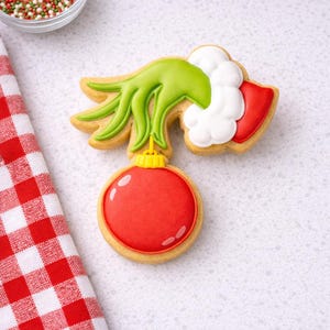May include: A decorated sugar cookie in the shape of a green hand holding a red ornament. The cookie has a yellow outline and is decorated with white icing and a red and white checkered napkin.
